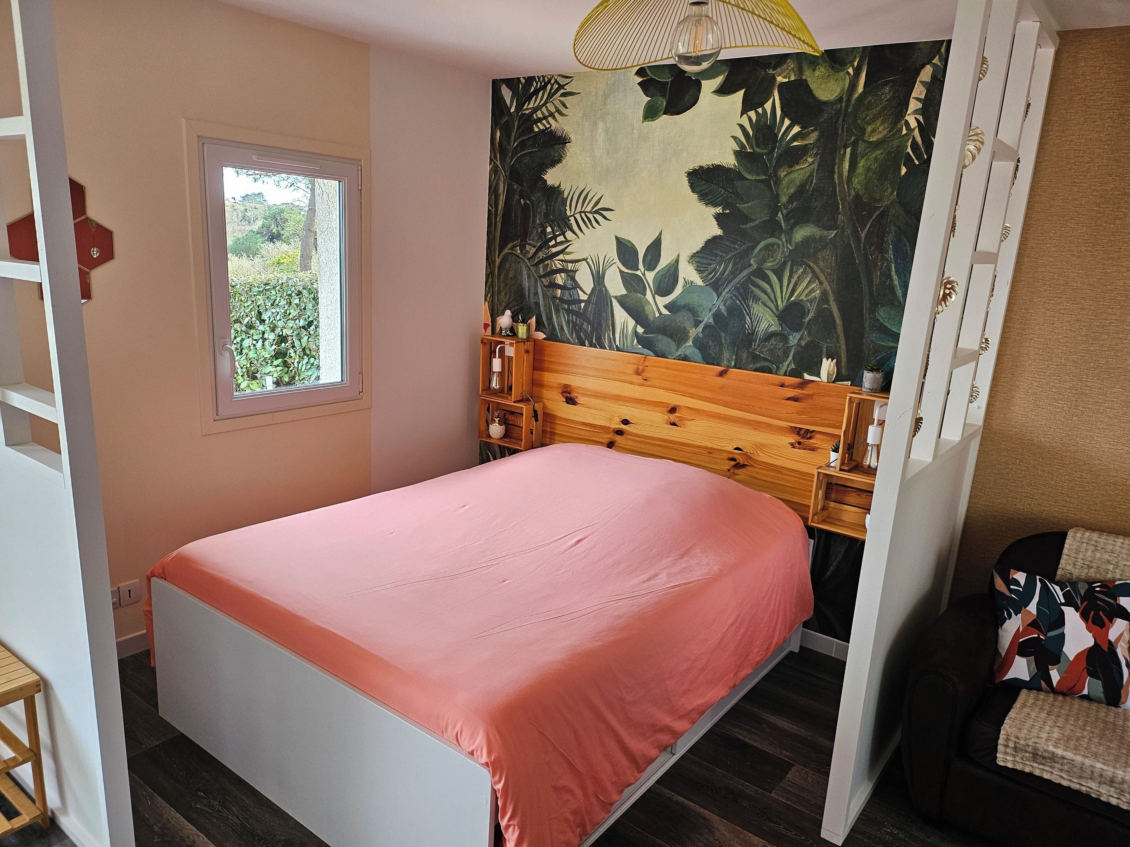 A bedroom with a bed, a window, a wooden headboard, and a wall with a tropical mural.