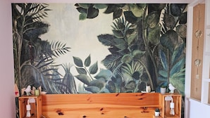 A bedroom with a wooden headboard, a pink bedspread, and a large tropical-themed tapestry on the wall. There are two wooden nightstands with small potted plants and decorative items.