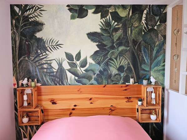 A bedroom with a wooden headboard, a pink bedspread, and a large tropical-themed tapestry on the wall. There are two wooden nightstands with small potted plants and decorative items.