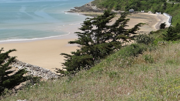 A coastal landscape with a sandy beach, a rocky shoreline, and a few houses on a hillside.