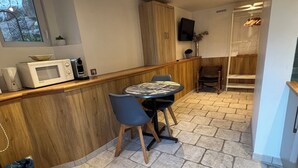 Uma cozinha compacta com micro-ondas, uma mesa pequena e cadeiras. Há um armário de madeira sobre o balcão e uma televisão fixada na parede.