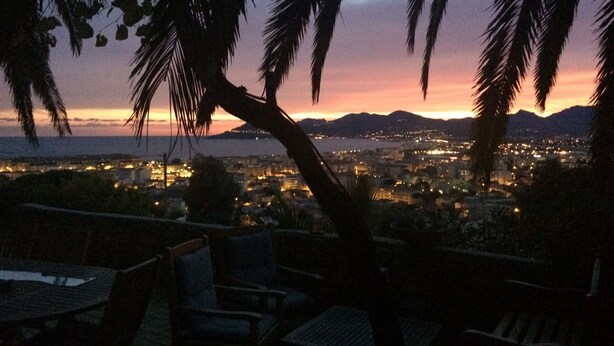 Vista do pôr do sol a partir de um terraço com palmeiras, com vista para uma paisagem urbana com edifícios e uma cordilheira distante.