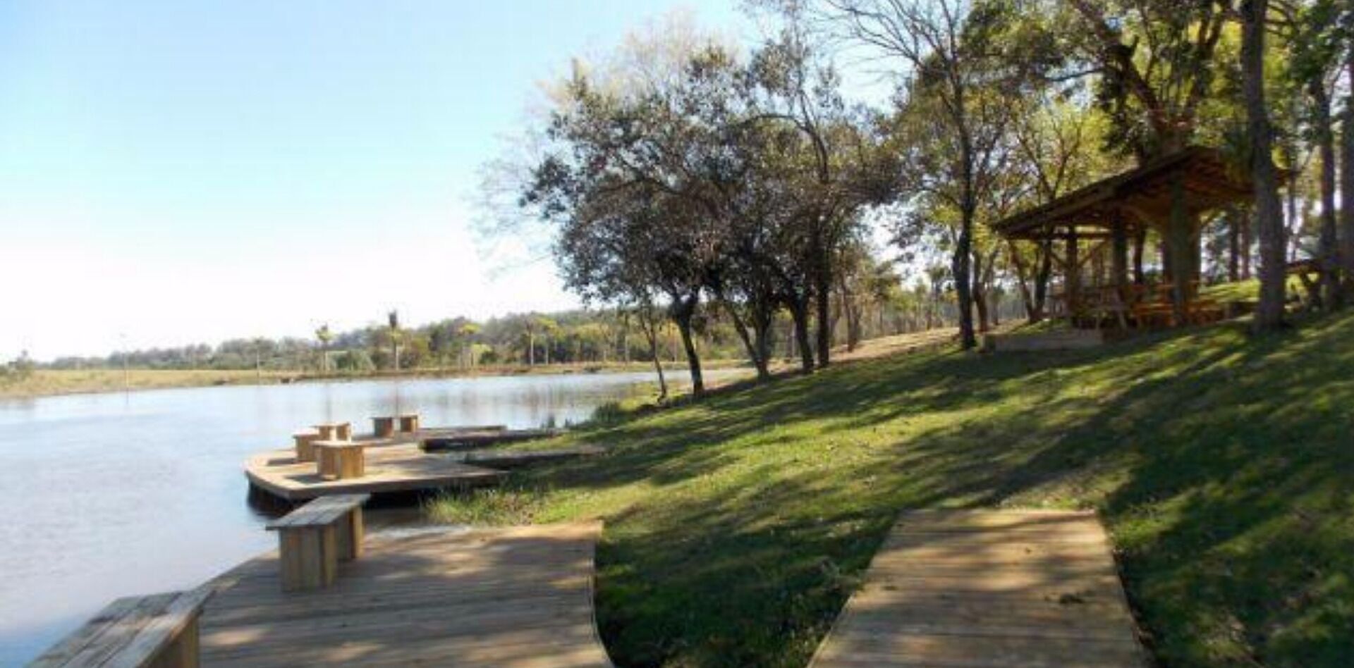 Une terrasse en bois au bord du lac, avec des tables de pique-nique, un espace de repos couvert et une pente herbeuse menant à un abri.