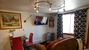 Ein gemütliches Wohnzimmer mit einem braunen Sofa, einem roten Sessel, einem Glastisch, einem an der Wand montierten Fernseher und einem gemusterten Vorhang.