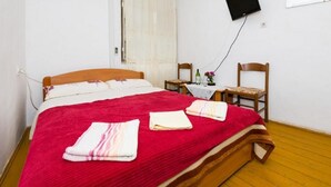 Een slaapkamer met een houten bed, een rode deken, twee handdoeken, een klein tafeltje met een fles en bloemen, en twee houten stoelen.