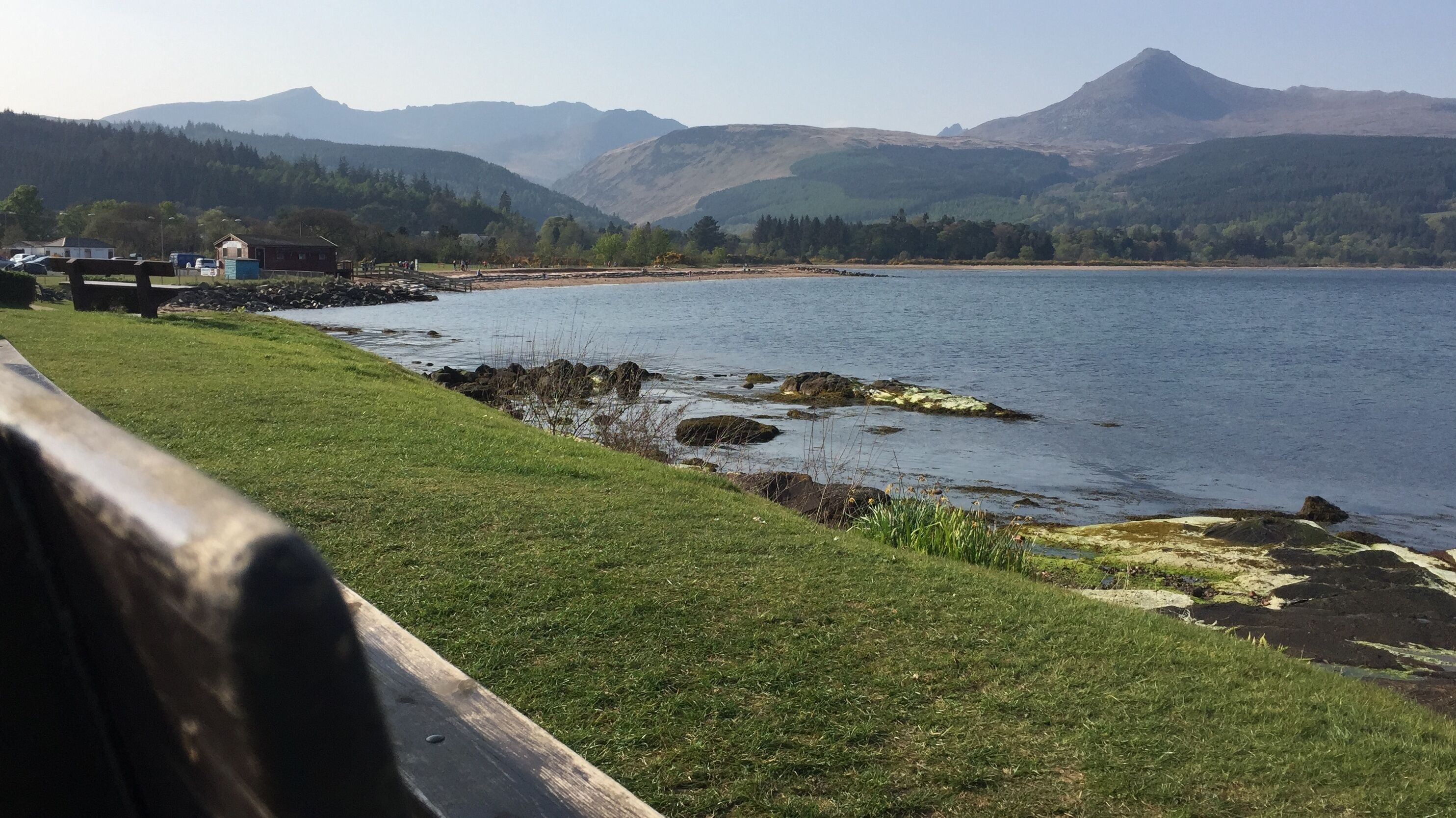 A coastal view with a grassy area, rocky shoreline, and mountains in the background.