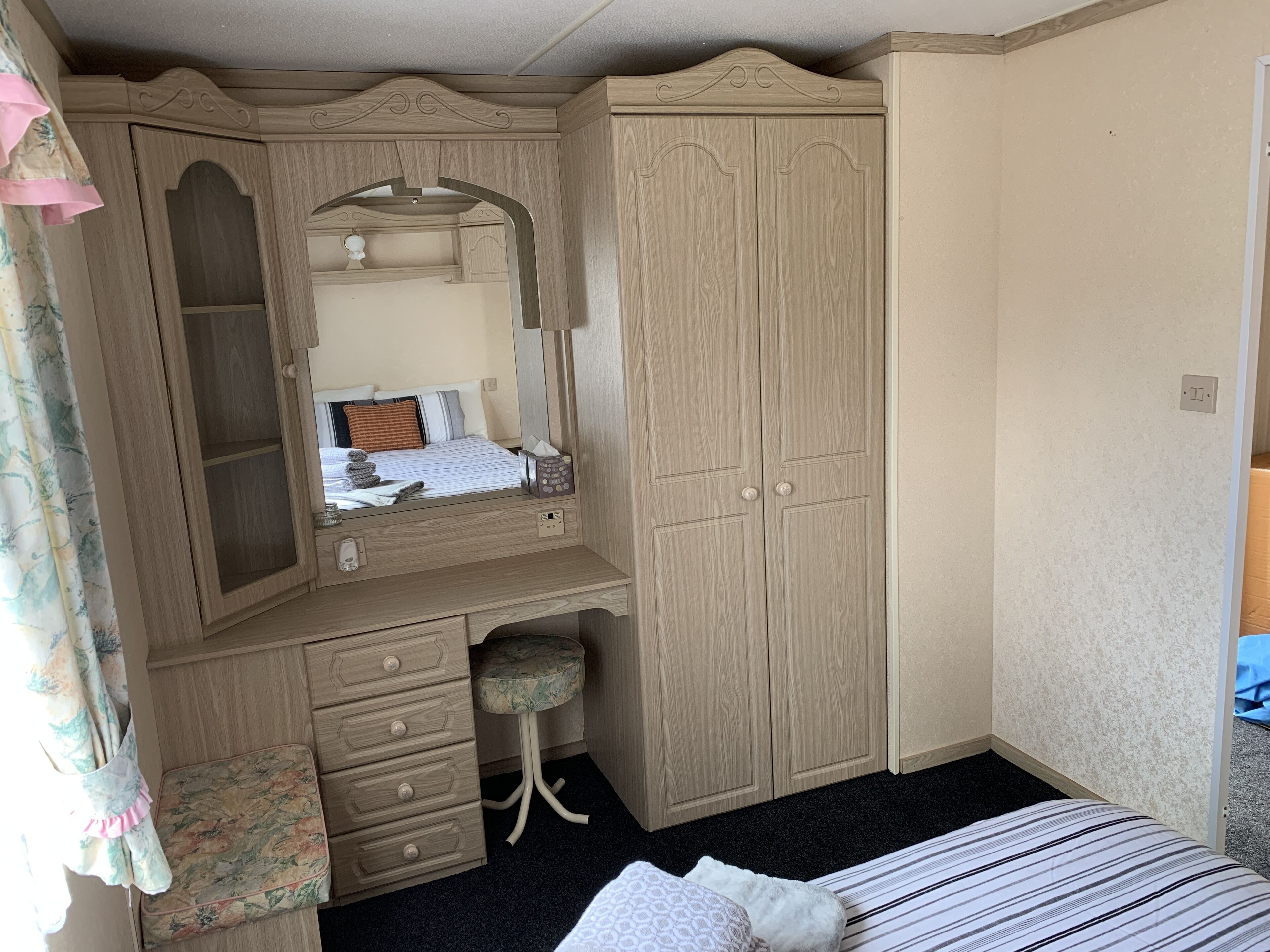 A bedroom with a wooden wardrobe, a mirror, a bed, and a small stool.