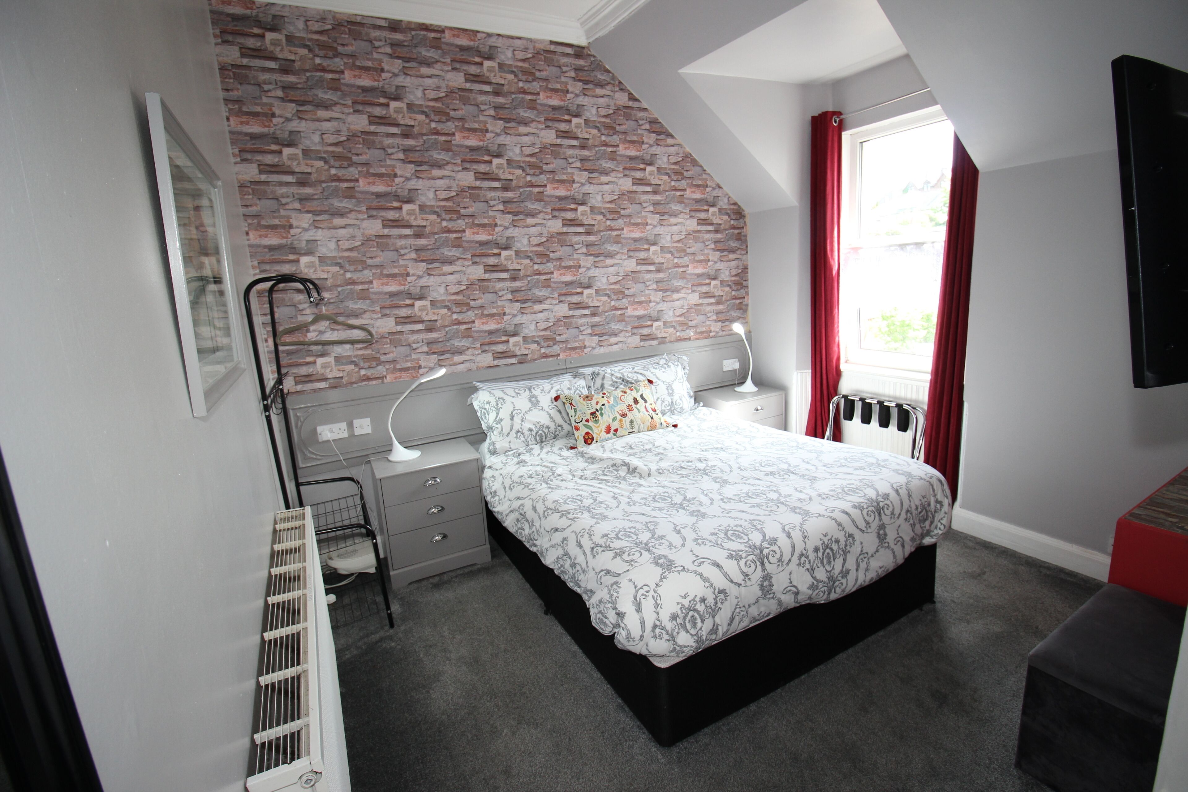 A bedroom with a brick-patterned wall, a bed with patterned bedding, a radiator, a window with red curtains, and a black bench.