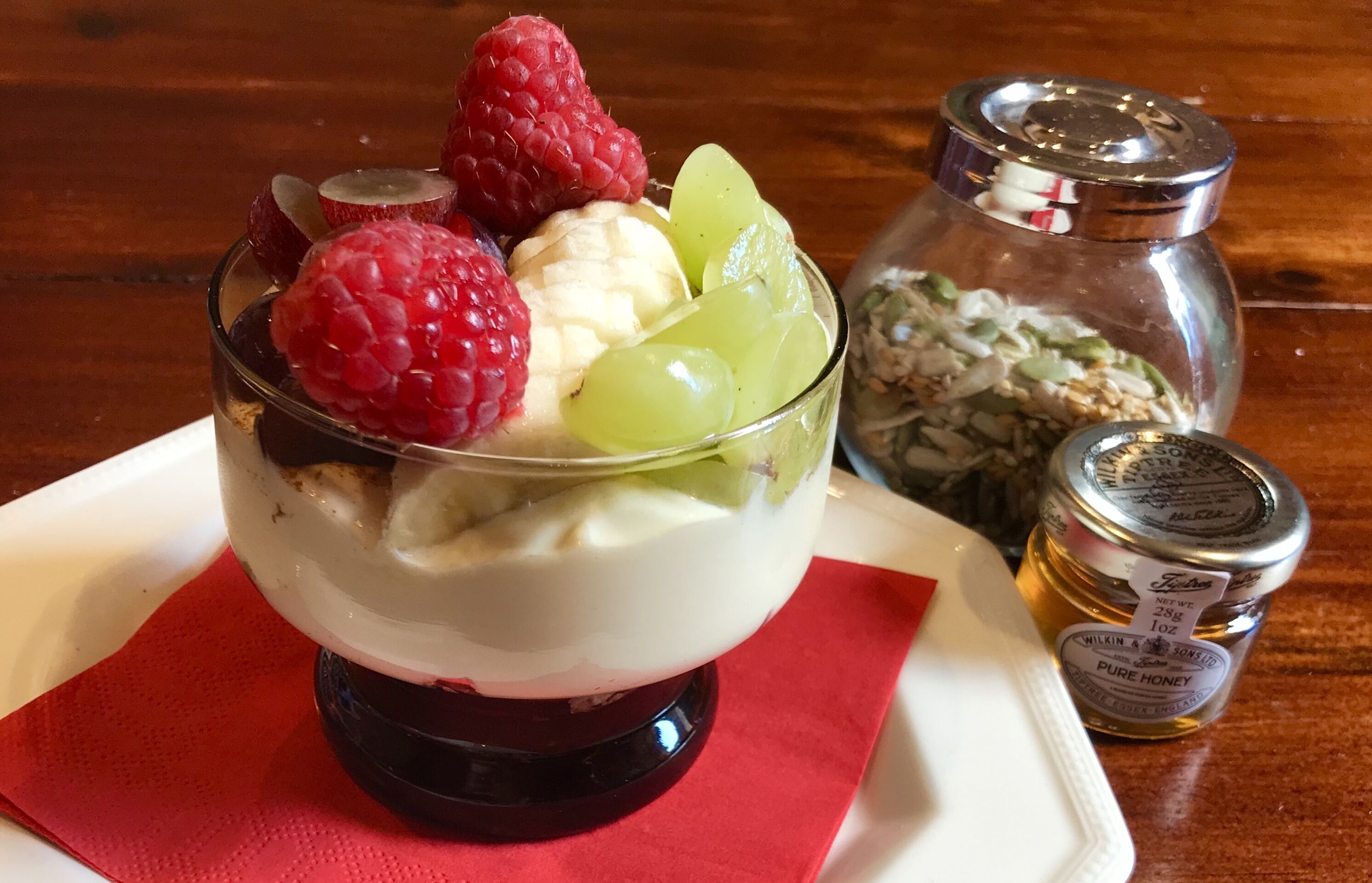 Un dessert composé de framboises, de raisins et de tranches de banane, présenté dans un plat en verre, accompagné d’un pot de miel et d’un récipient de flocons d’avoine.