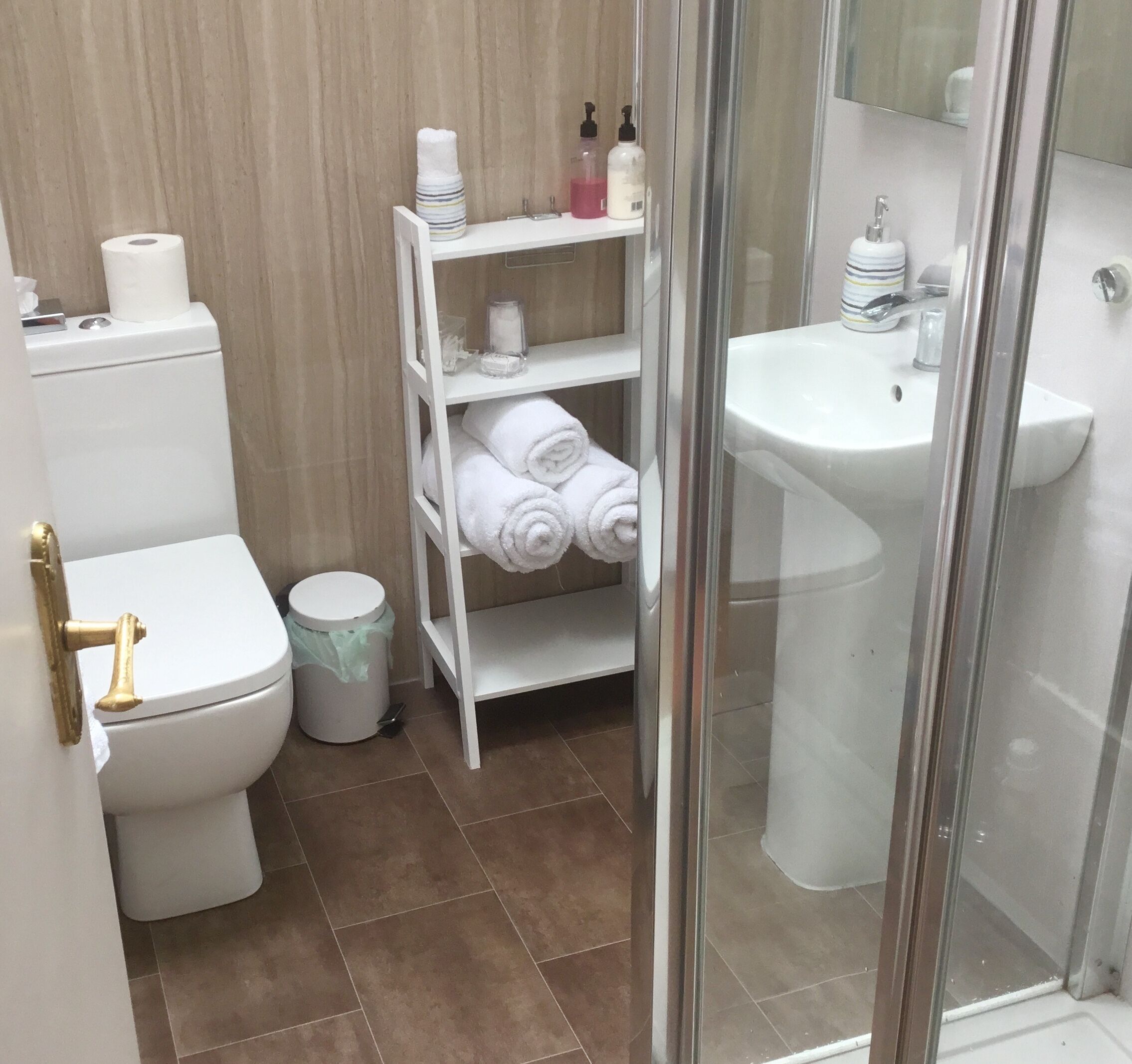 A bathroom with a toilet, a shelf with towels, a sink, and a trash bin.