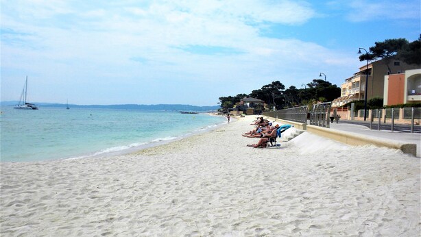 Ein Strand mit klarem türkisblauem Wasser, einem sandigen Ufer und einer Reihe von Gebäuden im Hintergrund.