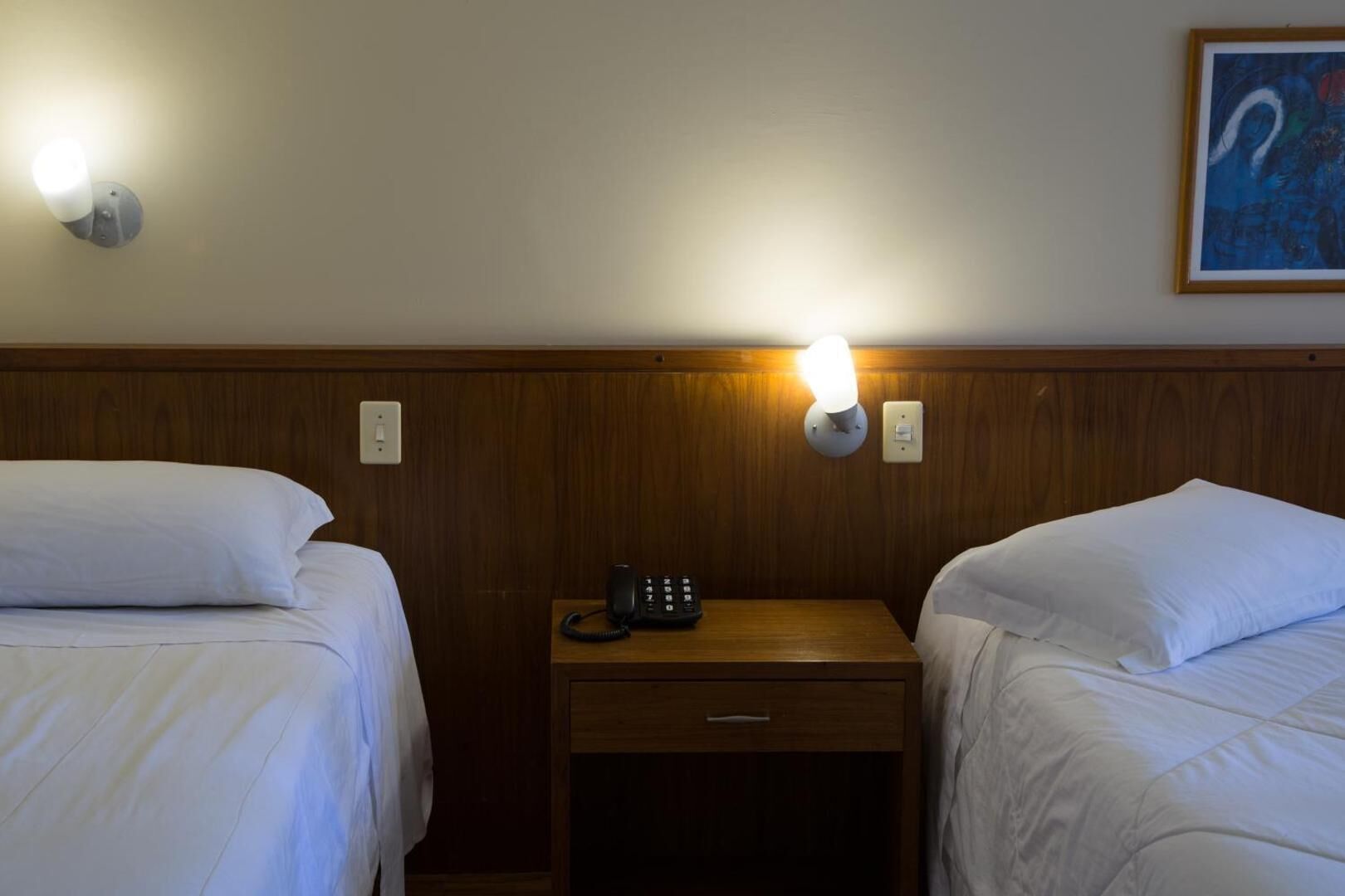 Habitación de hotel con dos camas individuales, cabecera de madera, mesita de noche con teléfono y lámpara fijada a la pared.