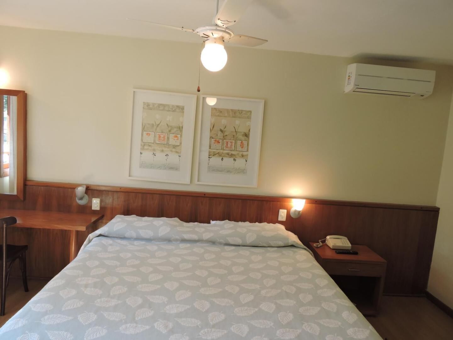 A hotel room with a bed, a desk, a chair, a nightstand, a phone, a ceiling fan, and two wall-mounted light fixtures.