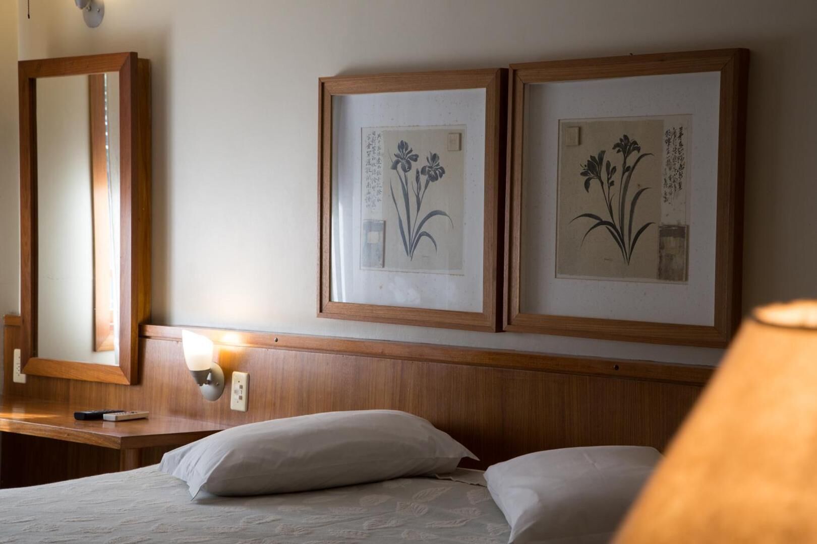 A hotel room with a wooden headboard, two framed botanical prints on the wall, a bedside table with a lamp, and a bed with white linens.