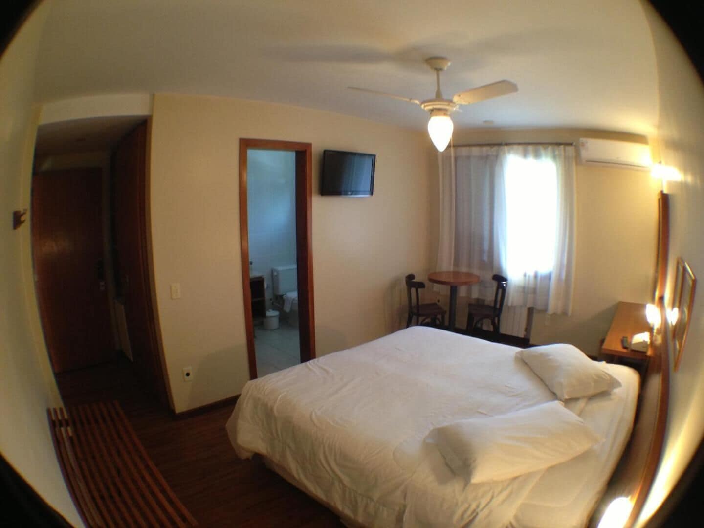 Habitación de hotel con cama, ventilador de techo, televisor, una pequeña mesa con sillas y ventana con cortinas.