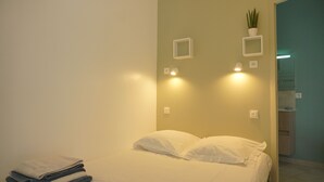 Ein ordentlich bezogenes Bett mit weißen Leinen in einem Zimmer mit hellgrünen Wänden. Zwei Wandleuchten und ein kleiner Blumentopf sind an der Wand angebracht.