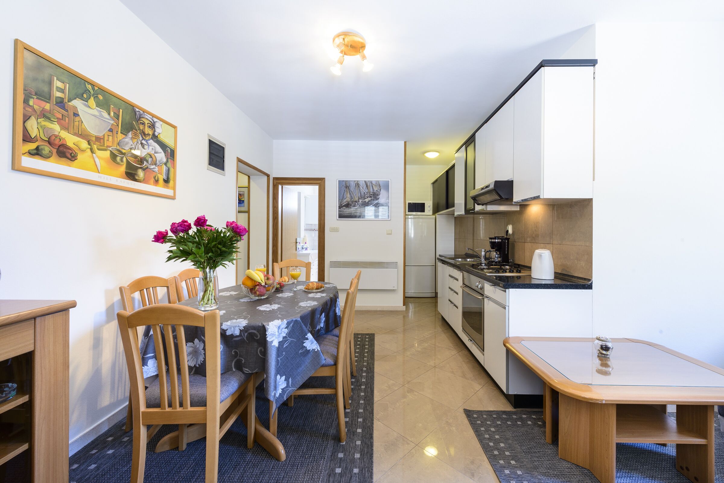 Una cocina compacta con electrodomésticos modernos, un comedor con mesa para dos personas y un cuadro en la pared.