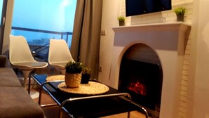 Una sala de estar moderna con chimenea, un televisor de pantalla plana montado sobre ella y un balcón con vistas al horizonte de la ciudad.