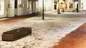 Une rue pavée recouverte de neige, bordée de bâtiments historiques, de bancs et où l’on aperçoit quelques piétons.