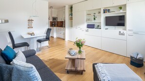 Un espace de vie moderne avec un canapé gris, une table basse en bois et un pouf gris. On y trouve également un coin cuisine avec des meubles blancs et un téléviseur à écran plat.