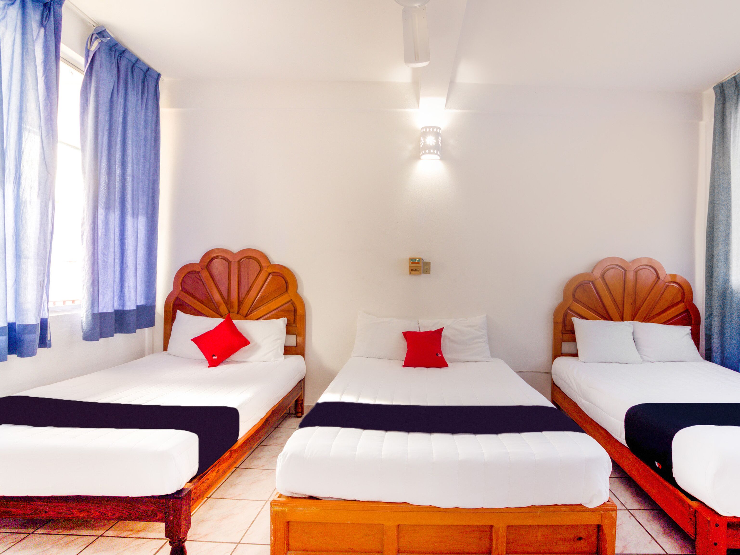 A hotel room with three single beds, each with a wooden headboard, white bedding, and a red pillow.