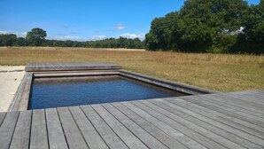 A rectangular swimming pool with a wooden deck surrounding it, set in a grassy field with trees in the background.