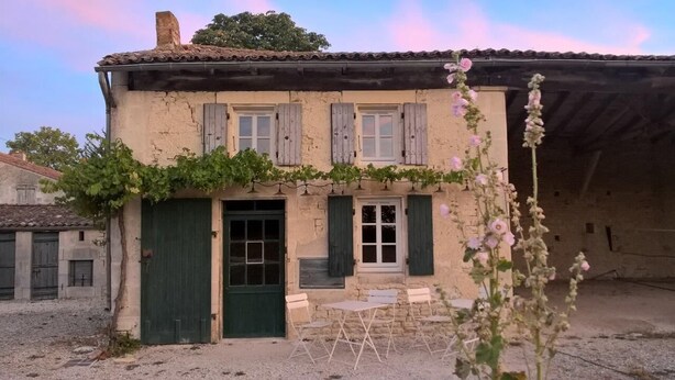 A rustic building with a green door, shuttered windows, and a small outdoor seating area.
