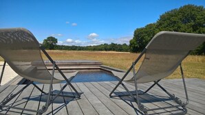 Two lounge chairs facing a small pool on a wooden deck with a view of a grassy field and trees.