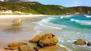 Una playa con aguas turquesas cristalinas, grandes rocas y colinas verdes al fondo.
