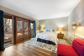 Un dormitorio con suelo de madera, una cama con edredón blanco, una mesita de noche con lámpara y vistas a través de un ventanal.