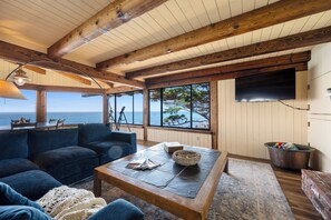 A cozy living room with a wooden ceiling, large windows overlooking the sea, a comfortable sofa, and a coffee table.
