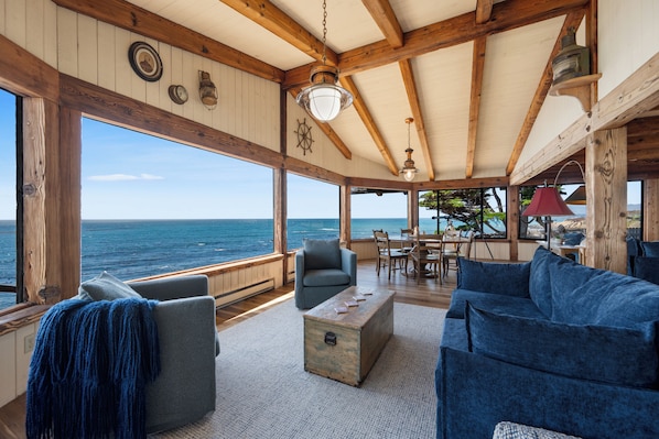 A cozy interior with a wooden ceiling, large windows overlooking the sea, a dining area, and a seating arrangement.