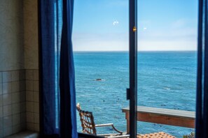 A view of the sea through a window with a chair and a wooden deck.