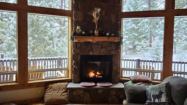 A cozy room with a stone fireplace, wooden ceiling, and large windows offering a view of snow-covered trees.