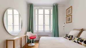 A bedroom with a bed, a round mirror, a chair, a window with curtains, and a radiator. - République - Furnished apartment - Downtown Lyon (Lyon)
