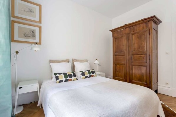 A bedroom with a bed, white walls, a wooden wardrobe, and two framed pictures.