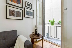 Ein Zimmer mit grauem Sofa, einem kleinen runden Tisch mit einer Vase und einem Balkon mit Blumentöpfen.