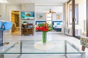 Un salon moderne avec une table basse en verre, un fauteuil bleu, un lit avec une literie bleue et un téléviseur fixé au mur.