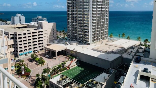 Un hôtel à plusieurs étages doté d’une piscine sur le toit et d’un court de tennis, offrant une vue sur une plage et l’océan.