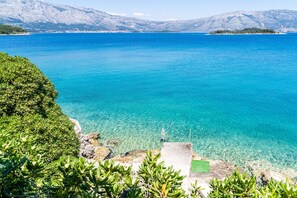 Un paisaje costero con aguas azules cristalinas, una costa rocosa y una cadena montañosa distante.