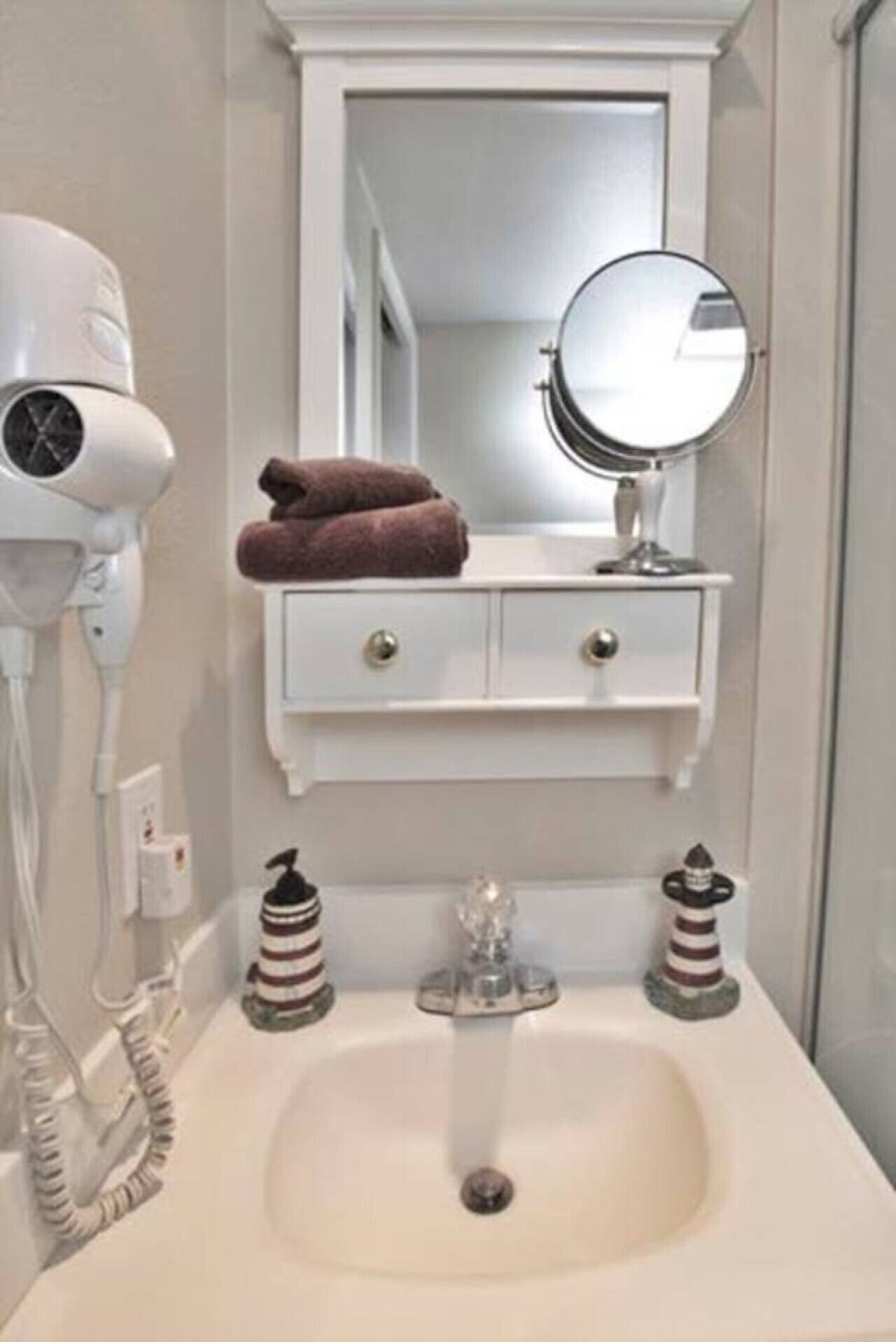 Une salle de bain avec un lavabo blanc, un sèche-cheveux, un miroir et deux bouteilles décoratives.