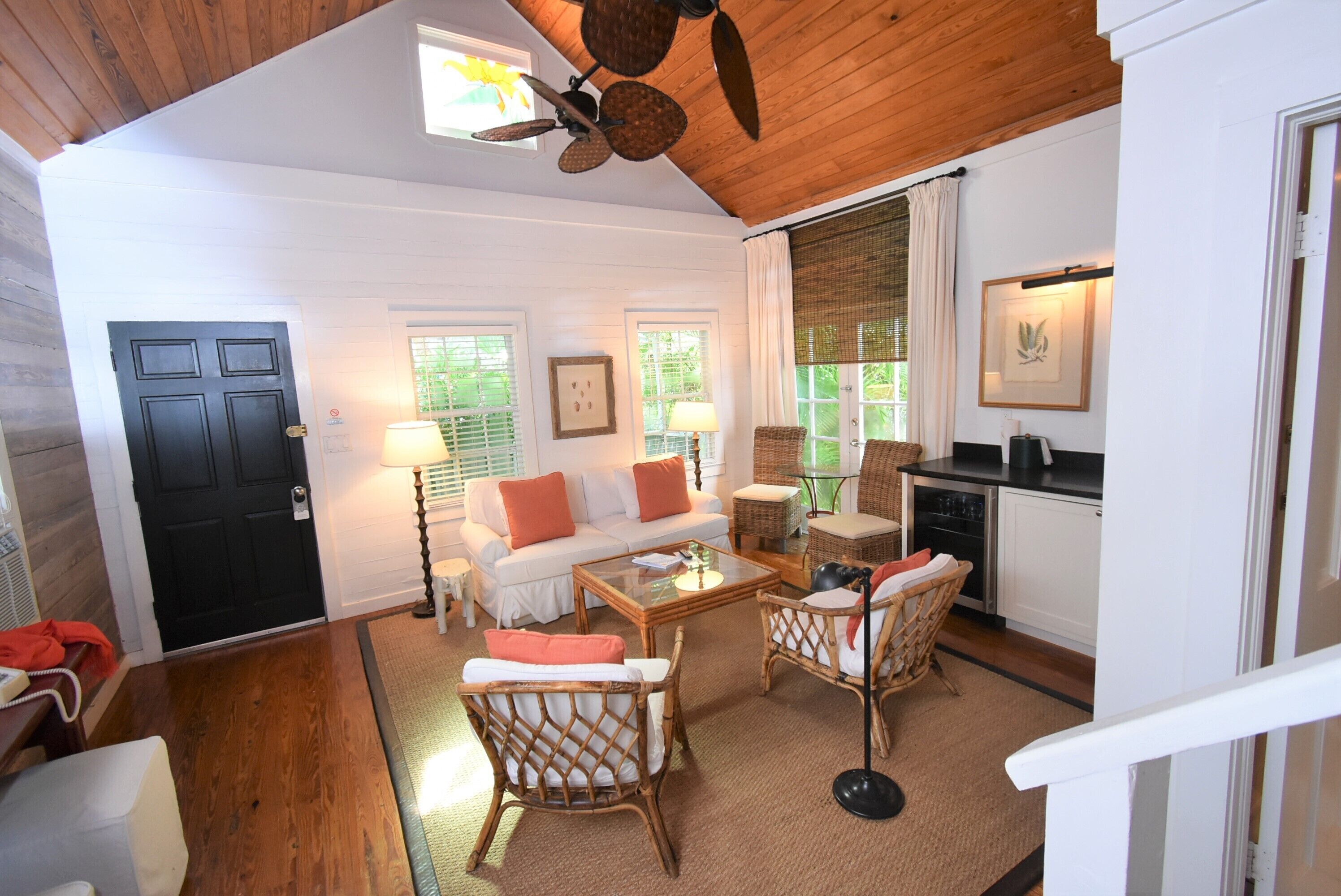 A cozy living room with a wooden ceiling, a black door, a white sofa with orange cushions, wicker chairs, a glass coffee table, and a kitchen area with a black countertop and white cabinets.