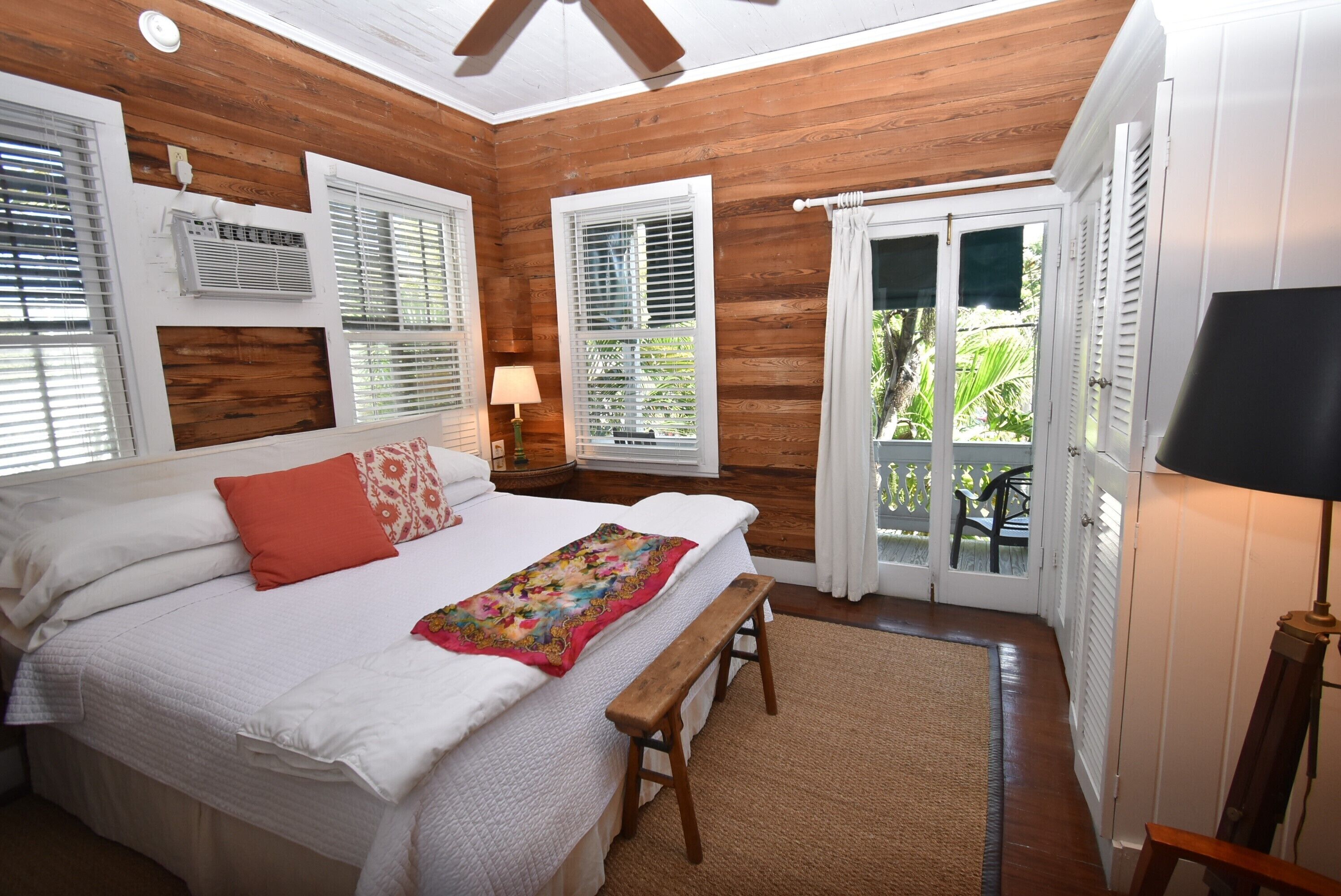 A bedroom with a large bed, wooden walls, white shutters, and a view of greenery outside.