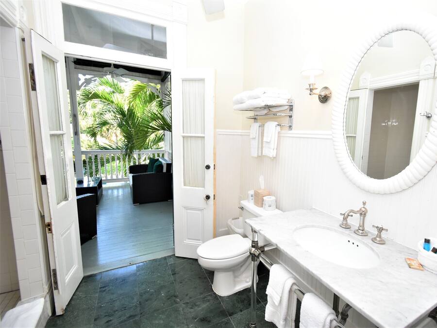 A bathroom with a white sink, a toilet, a large mirror, and a door leading to a balcony with a view of palm trees.