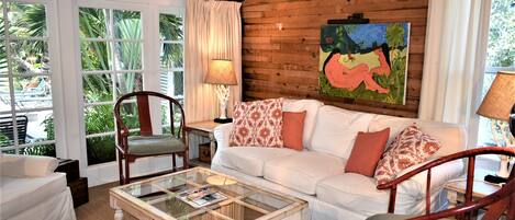 A cozy living room with a white sofa, patterned cushions, a glass coffee table, and wooden walls.