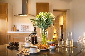 A breakfast setting with a coffee maker, a vase of flowers, croissants, and a bowl of fruit on a glass table.
