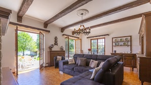 A spacious living room with a large sectional sofa, wooden beams on the ceiling, and a view of the outdoors through open doors.