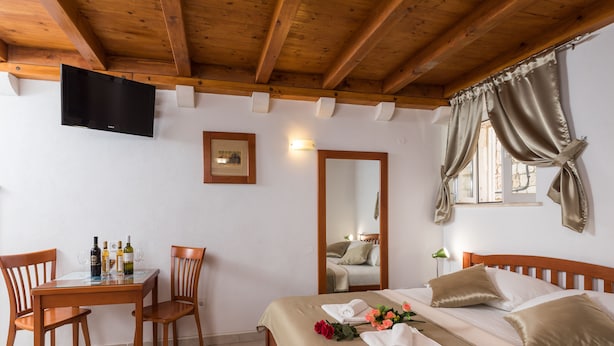 Ein Hotelzimmer mit Holzboden, einem Bett mit beigefarbener Bettwäsche, einem Holztisch mit Stühlen, einem an der Wand montierten Flachbildfernseher und einem Fenster mit Vorhängen.