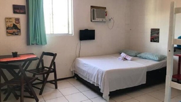 Habitació petita amb un llit individual, escriptori, cadires, finestra amb cortines, aire condicionat i decoració a les parets.