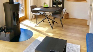 Un salon moderne avec un poêle à bois, une table à manger avec des chaises, un canapé bleu et une grande fenêtre donnant sur l’extérieur.